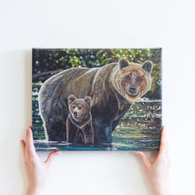 Load image into Gallery viewer, Two hands holding a small canvas reproduction of a painting. The painting features two grizzly bears, a mother, and her young cub crossing a river. They are standing in shiny green and turquoise water, and there is a blurry green background behind them.
