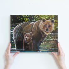 Load image into Gallery viewer, Two hands holding a small paper reproduction of a painting. The painting features two grizzly bears, a mother, and her young cub crossing a river. They are standing in shiny green and turquoise water, and there is a blurry green background behind them.
