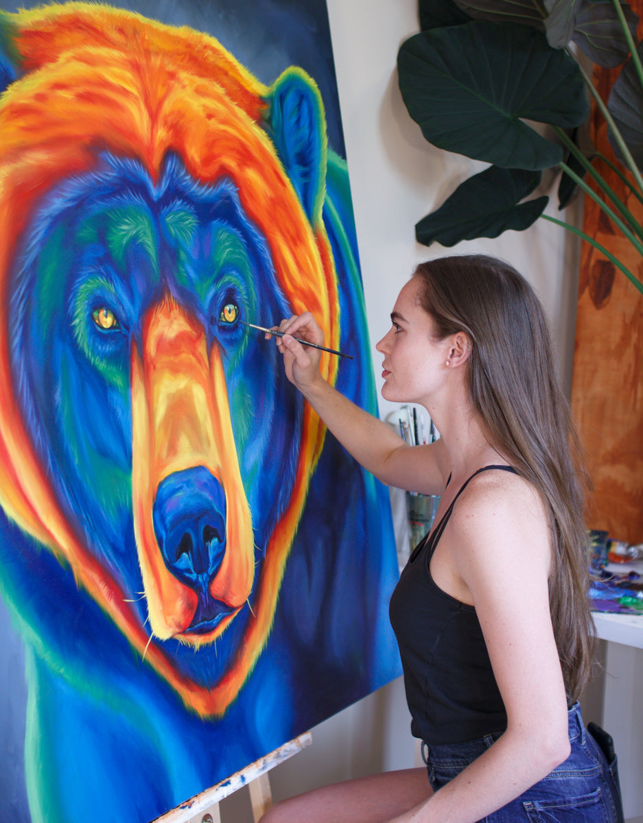 A brunette woman sits in front of a large painting of a rainbow bear. The painting she sits in front of is large, depicts a bear’s face, but the colours are altered so that the bear is painted in shades of red, orange, and yellow, green, blue, and purple.