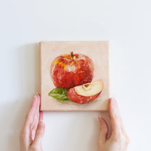 Load image into Gallery viewer, Two hands holding a small oil painting of an apple. The large, shiny apple is bright red on a peach coloured background. There is a slice of apple in the foreground with a green apple leaf to the left of it.
