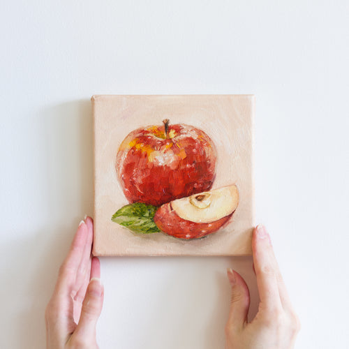 Two hands holding a small oil painting of an apple. The large, shiny apple is bright red on a peach coloured background. There is a slice of apple in the foreground with a green apple leaf to the left of it.