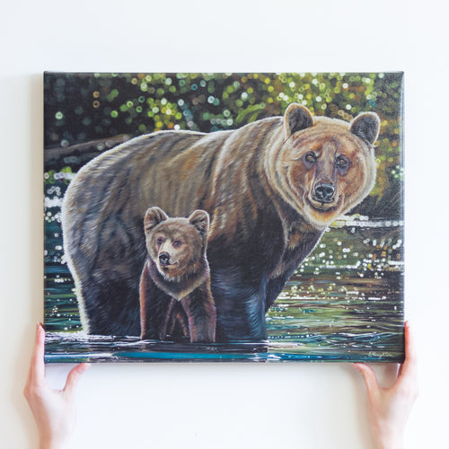 Two hands holding a large canvas reproduction of a painting. The painting features two grizzly bears, a mother, and her young cub crossing a river. They are standing in shiny green and turquoise water, and there is a blurry green background behind them.