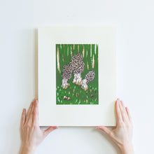 Load image into Gallery viewer, Two hands holding a small screenprint of three morel mushrooms on a dark green background. The print is made up of three colours, an emerald green, a light brown, and a dark brown. The print is small on the paper, with large white borders around it.
