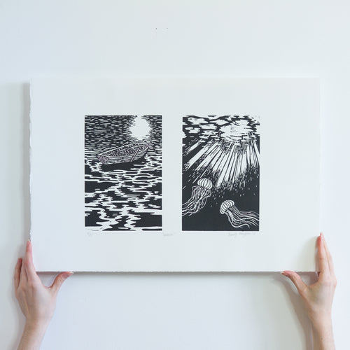 Two hands holding a black and white linocut print from the bottom. There are two images, the first of a boat sitting on the water at nighttime, reflecting the light of the moon. The second is of the light illuminating two jellyfish underneath the water.